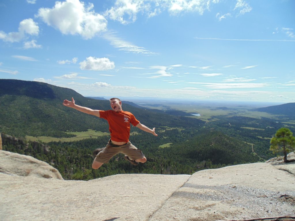 Teen boy on mountain top with blue sky in background jumping with excitement